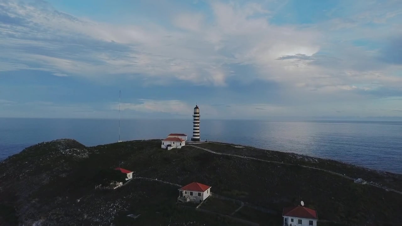 Farol de Abrolhos sull'Ilha de Santa Bárbara, Parco Nazionale Marino di Abrolhos, Caravelas, Bahia, Brasile, con faro a strisce, case del personale e cresta rocciosa sotto un cielo nuvoloso, ripresa lenta in avanti con drone.