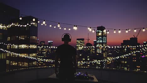 silhouetted professional dj performing on urban rooftop during sunset, overlooking illuminated city skyline with glowing string lights creating vibrant nightlife atmosphere