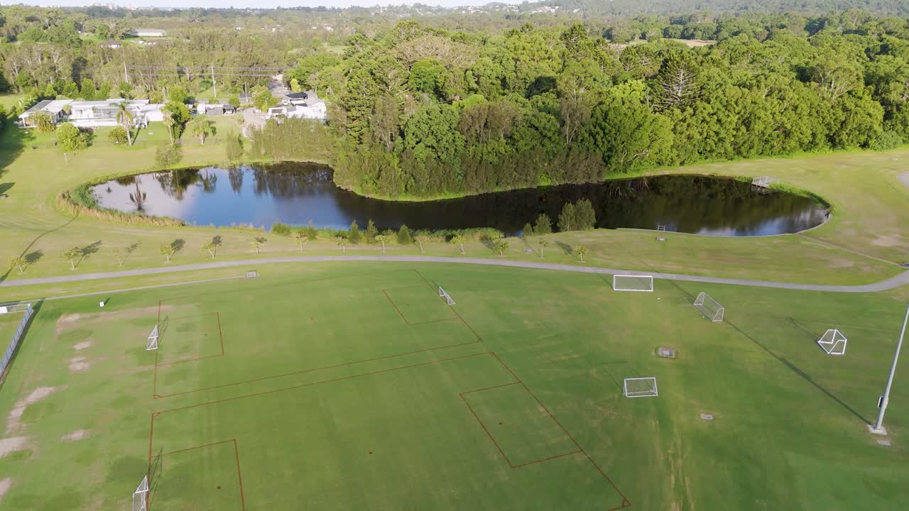Drone captures expansive soccer fields and lush greenery under bright daylight. The serene environment includes a reflective pond and distant trees