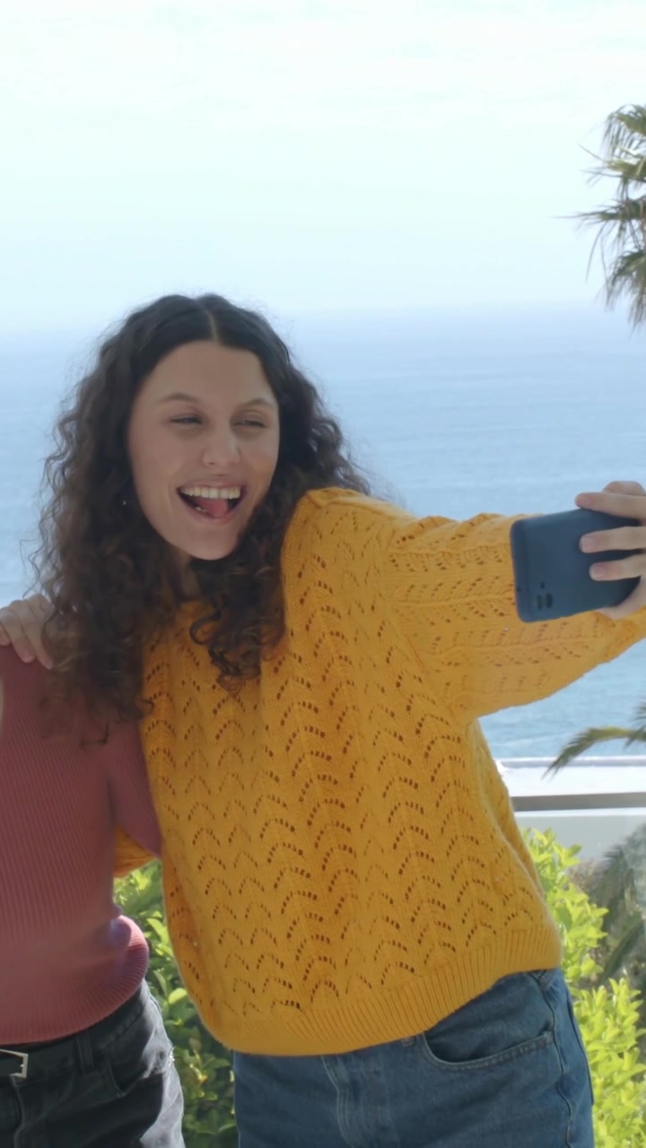 Video verticale: Donna in top color senape si appoggia a un'amica che tiene un telefono, scattano selfie sul balcone.