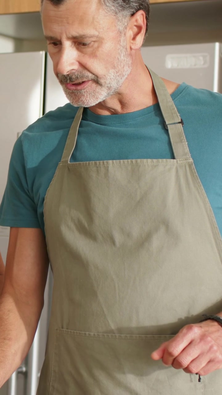 Video verticale: Uomo con grembiule beige che cucina in cucina, pizzicando il cibo, con un avambraccio che si intravede fuori campo.