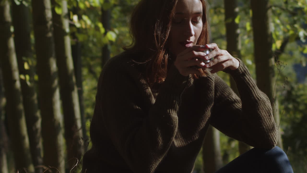 una joven sentada en la hierba en medio del bosque disfrutando de una taza de café caliente de un termo