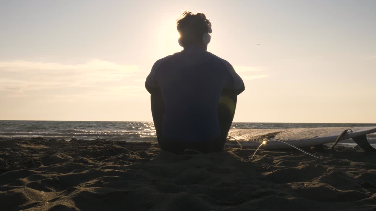 joven surfista en traje de buceo sentado con una tabla de surf acostado en la arena en la playa escuchando música bailando mirando el océano al atardecer