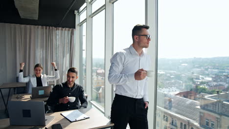 lady sitting at desk in front of laptop has had a successful deal and is very excited. man in white shirt drinks coffee standing at window. sunny office backdrop.