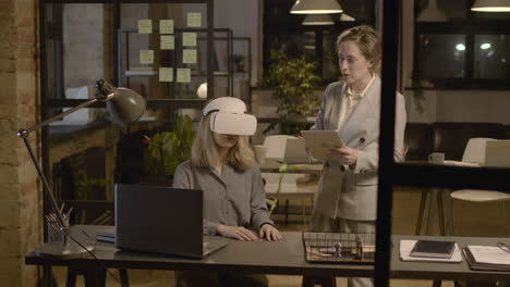 woman emplooyee at desk in the office wearing virtual reality glasses while her coworker is controlling her with tablet 2