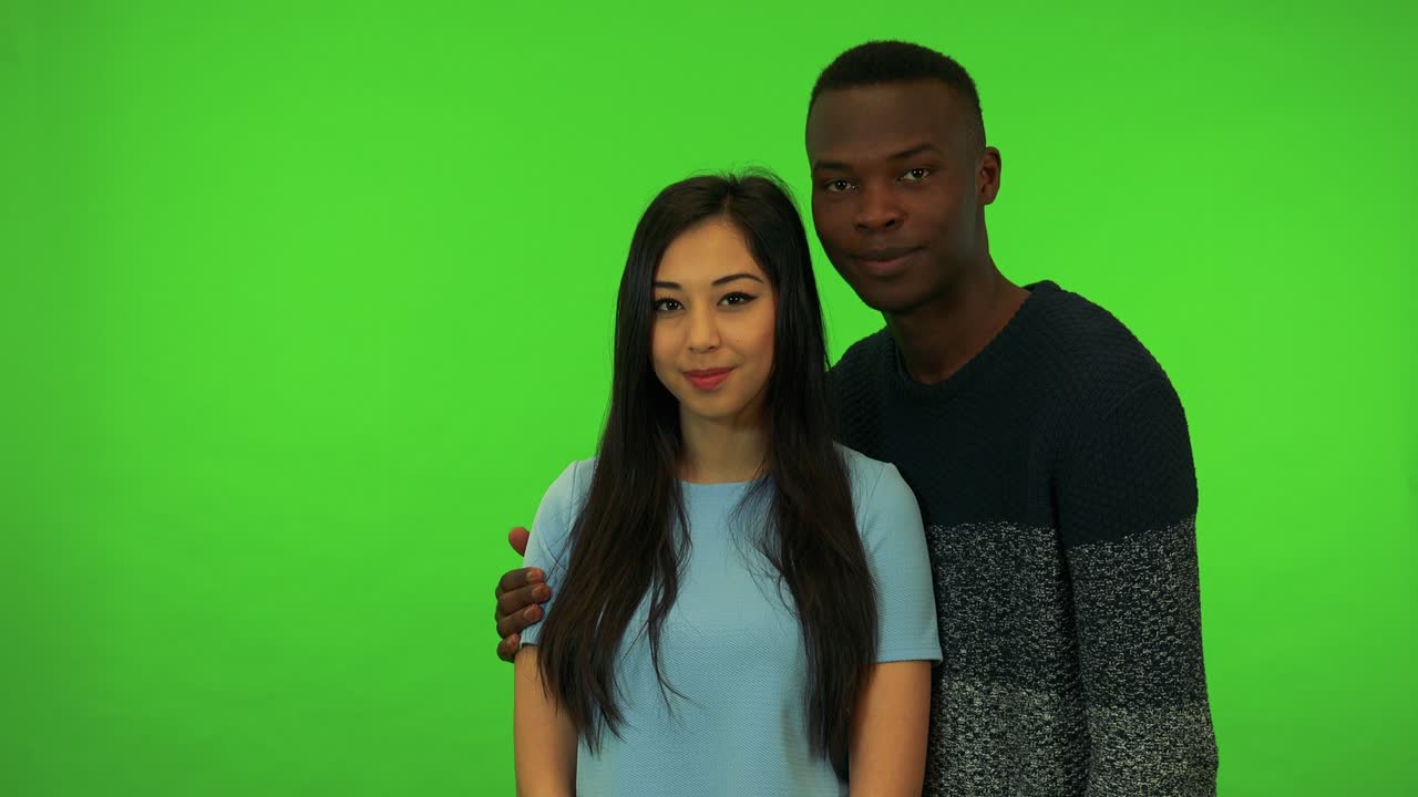 una joven asiática y un joven negro se dan una palmada, él envuelve su brazo alrededor de sus hombros y sonríen a la cámara - estudio de pantalla verde