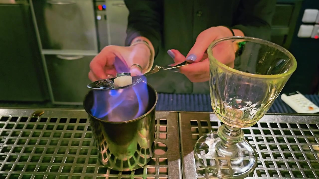 Preparazione tradizionale di assenzio in un bar buio, con zolletta di zucchero in fiamme, fiamme blu, tazza di metallo, bicchiere decorato, mani del barista e un'atmosfera di mixology drammatica all'interno