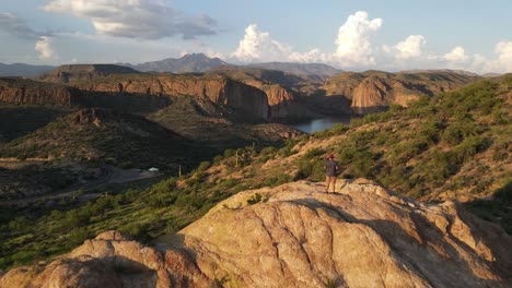 Drone-flying-toward-a-person-standing-on-the-edge-of-a-mountain-at-golden-hour-looking-down,-mountain-and-lake-in-the-background-with-light-blue-sky-and-patches-of-large-white-clouds-in-Arizona