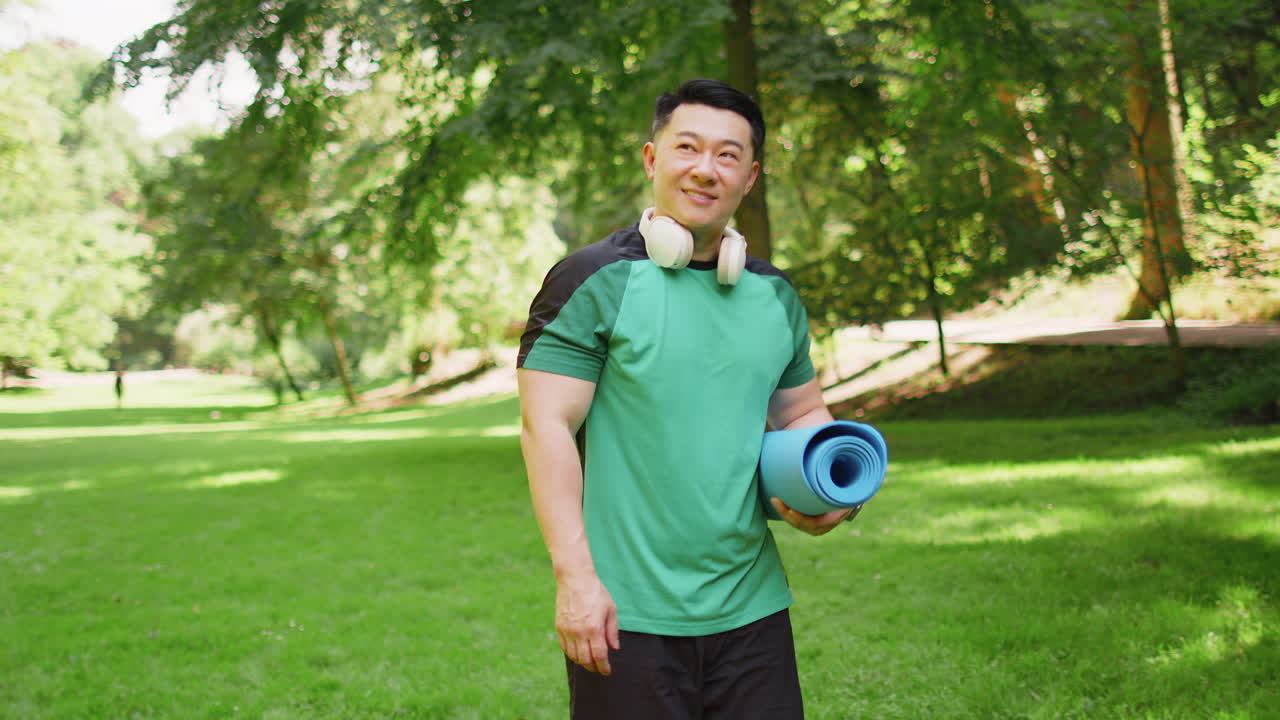 Athletic young man walking with rolled yoga mat to do fitness stretching workout exercises in park