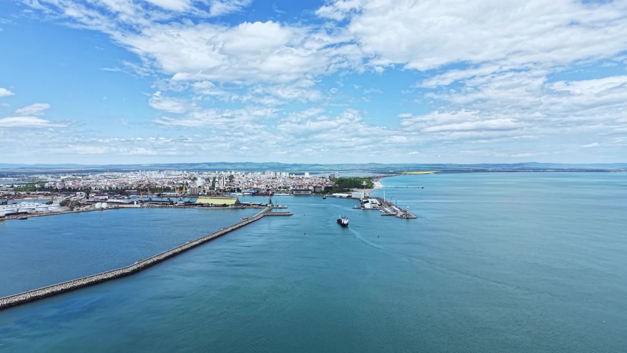Vista di una città costiera sull'acqua con navi e un porto durante il giorno, con nuvole nel cielo e mare calmo.