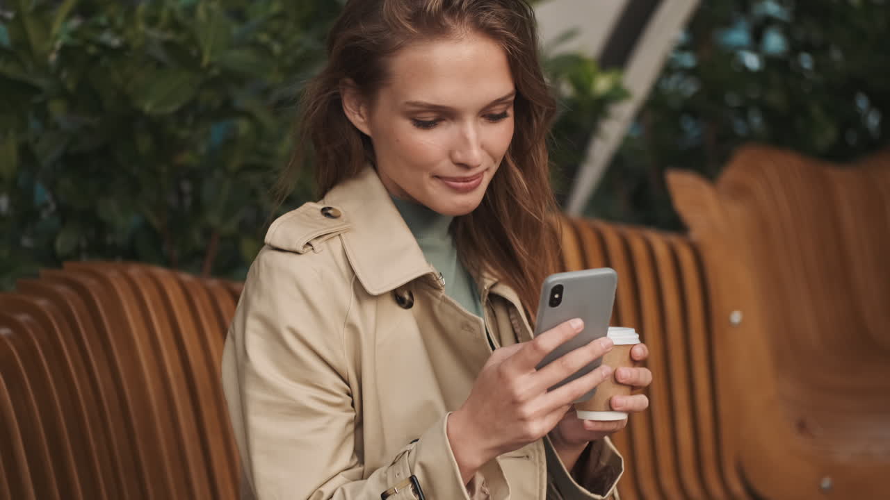 Caucasian female student using smartphone and drinking coffee outdoors.