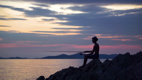 Junger-Mann-Mit-Hut,-Der-Auf-Den-Felsen-In-Der-Nähe-Des-Meeres-Sitzt-Und-Versucht,-Während-Des-Sonnenuntergangs-Fische-Zu-Fangen---Weitschuss