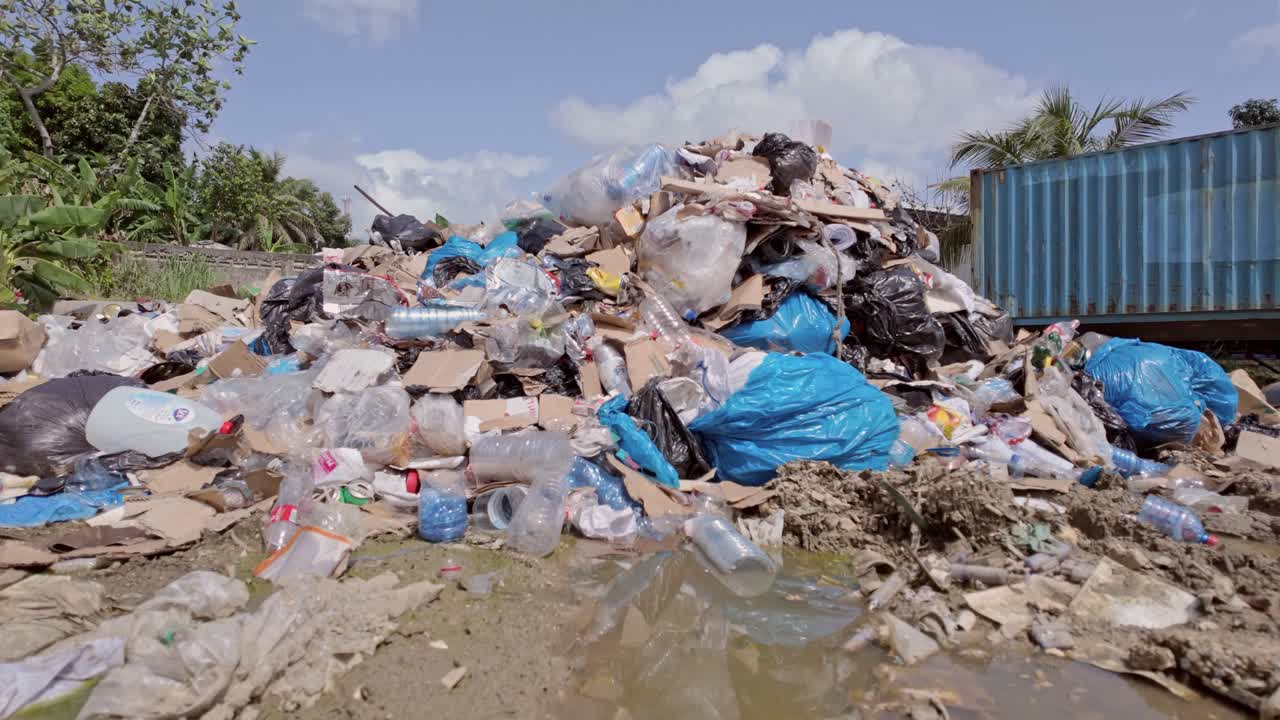 vista hacia arriba desde el nivel del suelo de un vertedero de basura en un día soleado, con agua sucia, plásticos desperdiciados, un contenedor abandonado, árboles y nubes en el cielo
