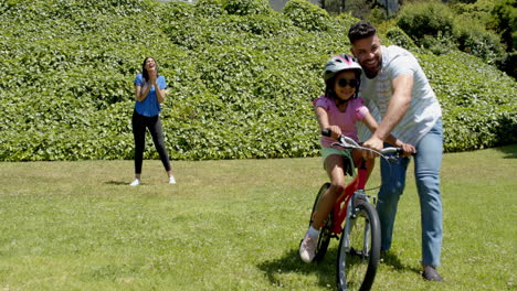 father teaching daughter to ride bicycle while mother cheers in sunny park