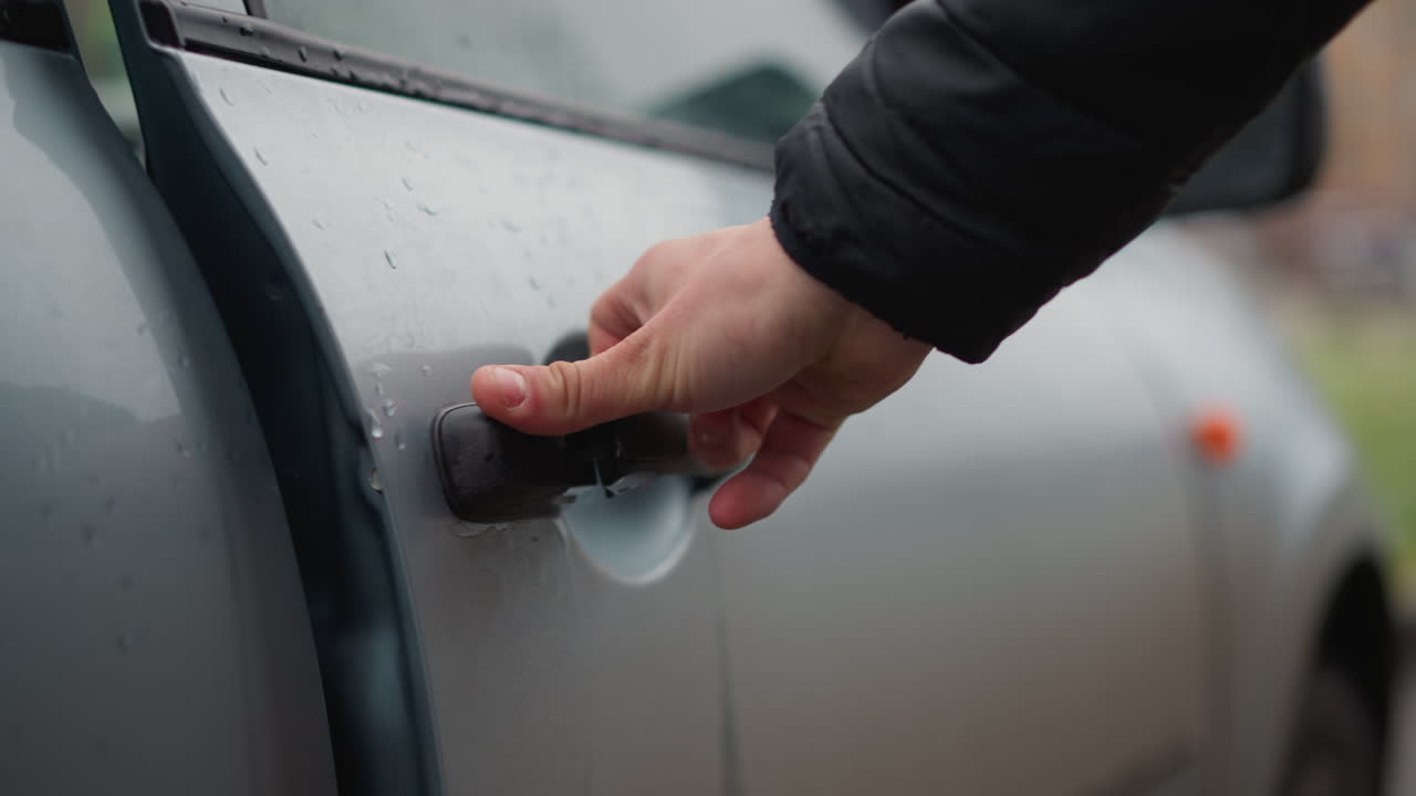 Close up hand of young adult opening car door handle on silver vehicle with water droplets showing winter environment and cold weather during urban commute scene on rainy pavement near parked cars