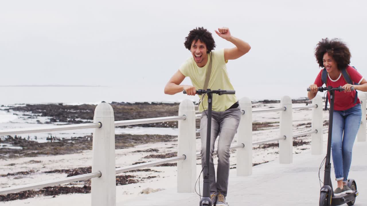 pareja afroamericana sonriendo mientras conducen scooters juntos en el paseo marítimo cerca de la playa