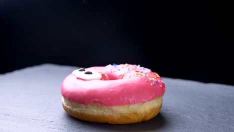 close-up circling around shot of delicious glazed pink donut with multicolored chips and eyes spinning slowly on gray table background.