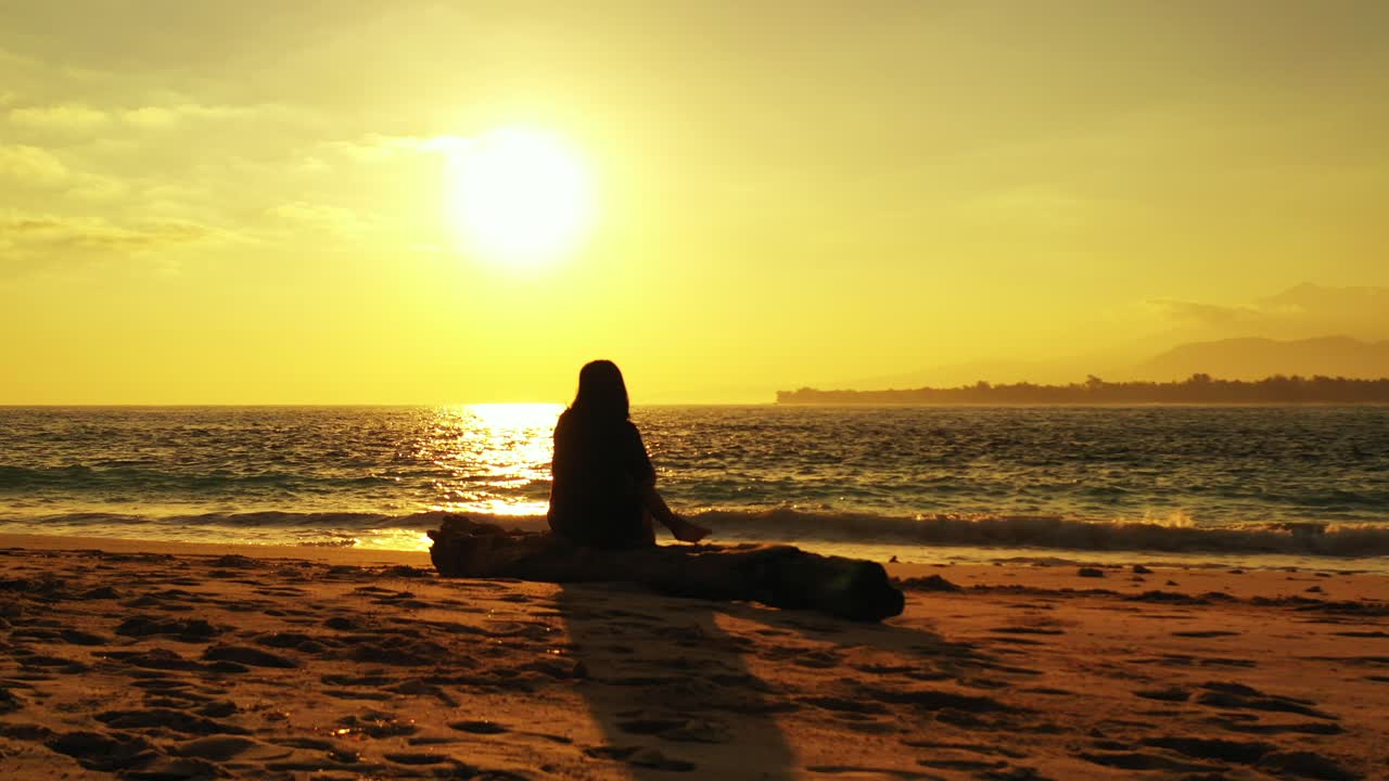 chica disfrutando de una hermosa puesta de sol con un cielo naranja amarillo reflejado en la superficie del mar sentada en una playa exótica en indonesia