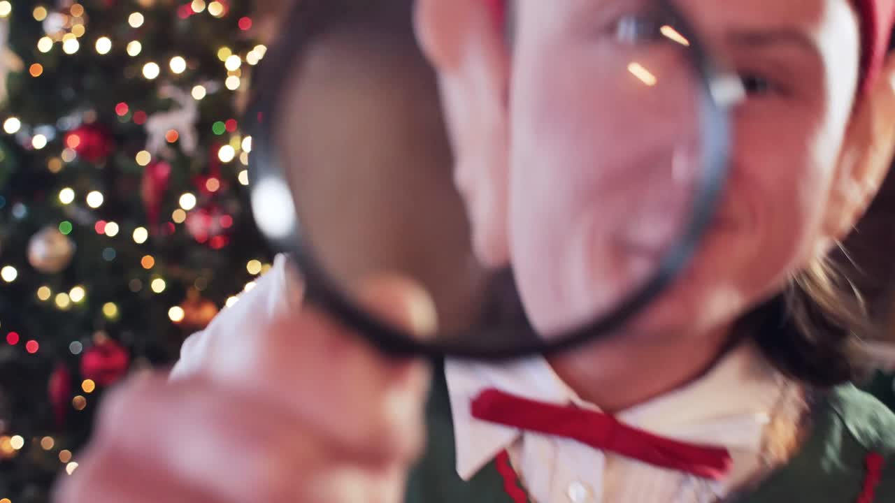 Elf looks into the camera through a magnifying glass, showing teeth. Christmas tree in the background. Close-up