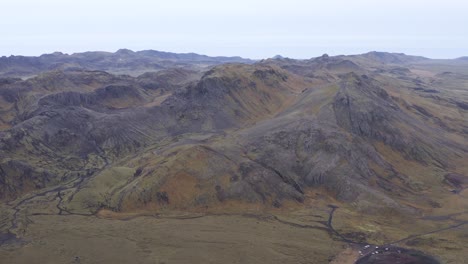 Volcanic-mountains-rise-high-above-grass-plains-in-Icelandic-landscape,-aerial