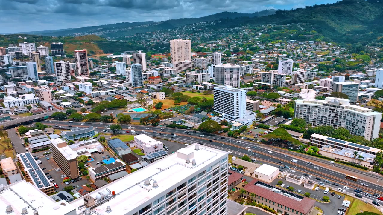 Traffico vivace sulle strade di Honolulu durante il giorno. Il drone si solleva sopra il panorama della città rivelando la vista sulle montagne