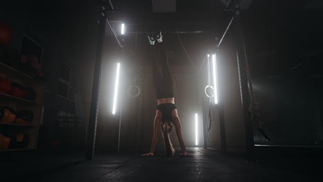 A-strong-young-athletic-woman-goes-upside-down-on-her-hands-in-the-gym.-A-woman-is-fit-on-her-hands-in-a-dark-gym.-Training-at-least-upside-down