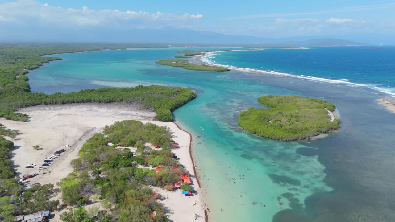 Parking cars of tourist at Playa Caobita Beach with clear water bay. Aerial top down shot. Sunny day in Dominican Republic. Green islands along coastline. Wide shot.