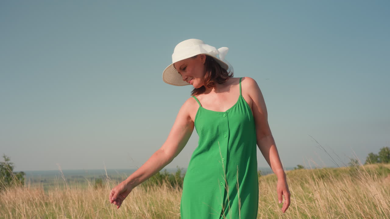 stylish lady in green dress and white sun hat standing in tall golden grass bends forward to gently pull out single grass blade while gazing down in serene summertime field