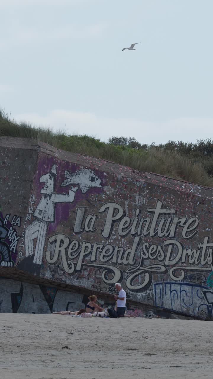 Family sets up leisure spot beside colorful graffiti bunker on sandy coastal dunes, natural daylight
