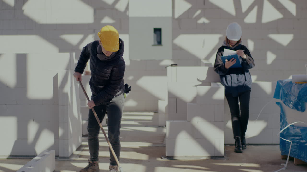 Young female worker brooming by supervisor holding clipboard near male coworker examining wall with level at construction site
