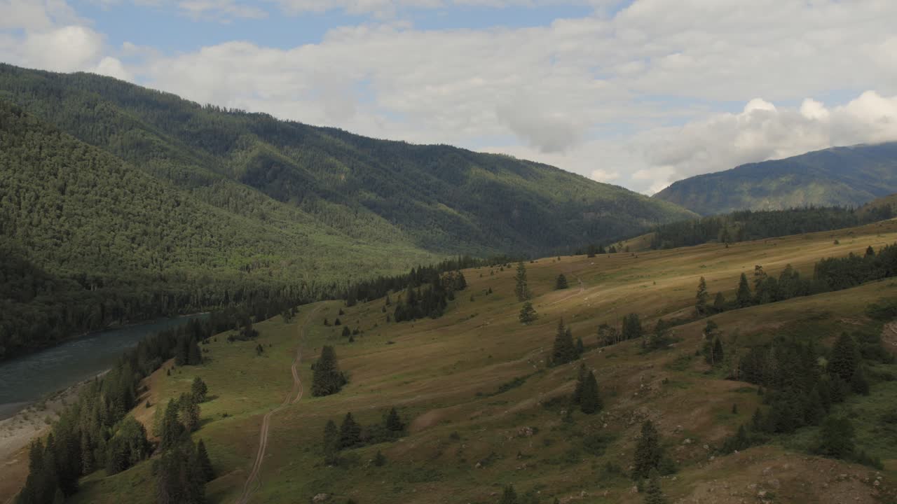 Ampio panorama aereo della Valle di Ak-Kem nei Monti Altai, Russia, con pendii boscosi, prati aperti e sentieri tortuosi sotto un cielo estivo nuvoloso