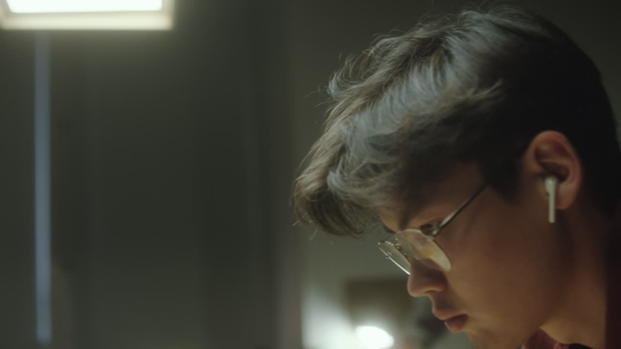 Focused Young Man with Earbuds and Glasses in a Dimly Lit Room