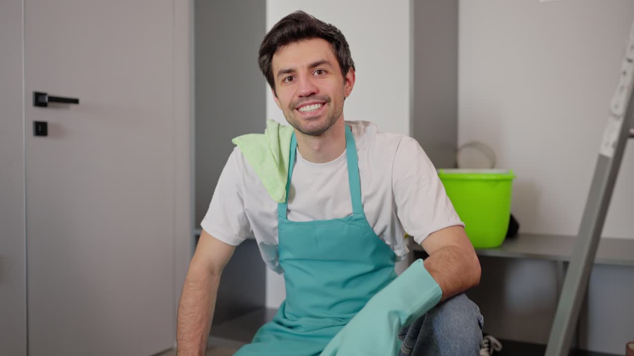 retrato de un seguro feliz hombre moreno limpiador en un delantal azul en una camiseta blanca que trabaja para una compañía de limpieza de llamada ahora en un apartamento moderno