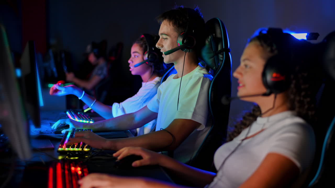 Multiracial group of teens in headsets playing video games in video game club with blue and red illumination. Keyboards and mice with illumination