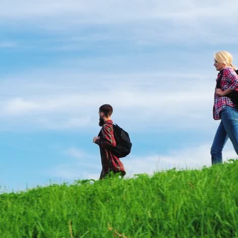 A-Couple-Of-Tourists-With-Backpacks-Are-Walking-Along-The-Crest-Of-A-Large-Green-Hill-8