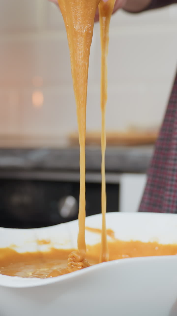 Close-up of woman holding whisk with thick caramel-colored pap stretching as it drips back into white bowl, kitchen background with blurred cookbook, candle, and flour-dusted countertop