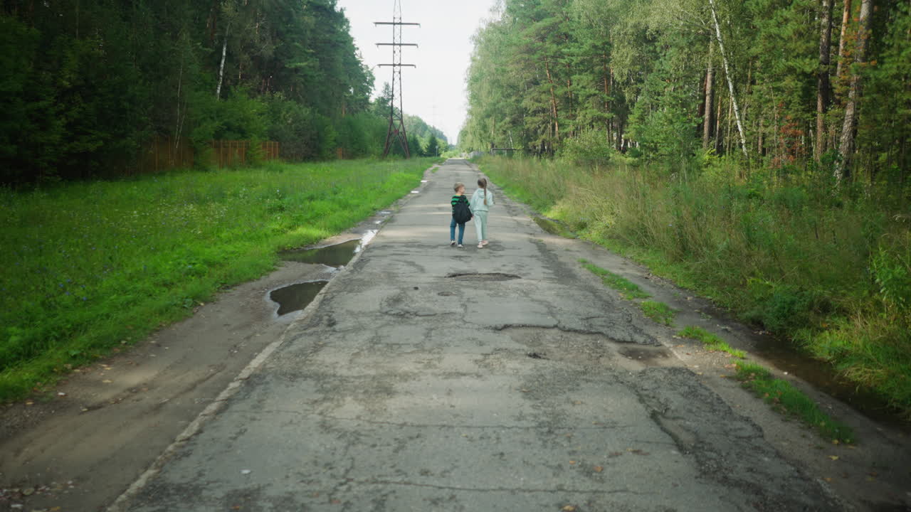 Rear view of young boy adjusting backpack while walking with sister along worn tarred road flanked by trees and puddles, under utility poles in peaceful outdoor setting surrounded by lush greenery
