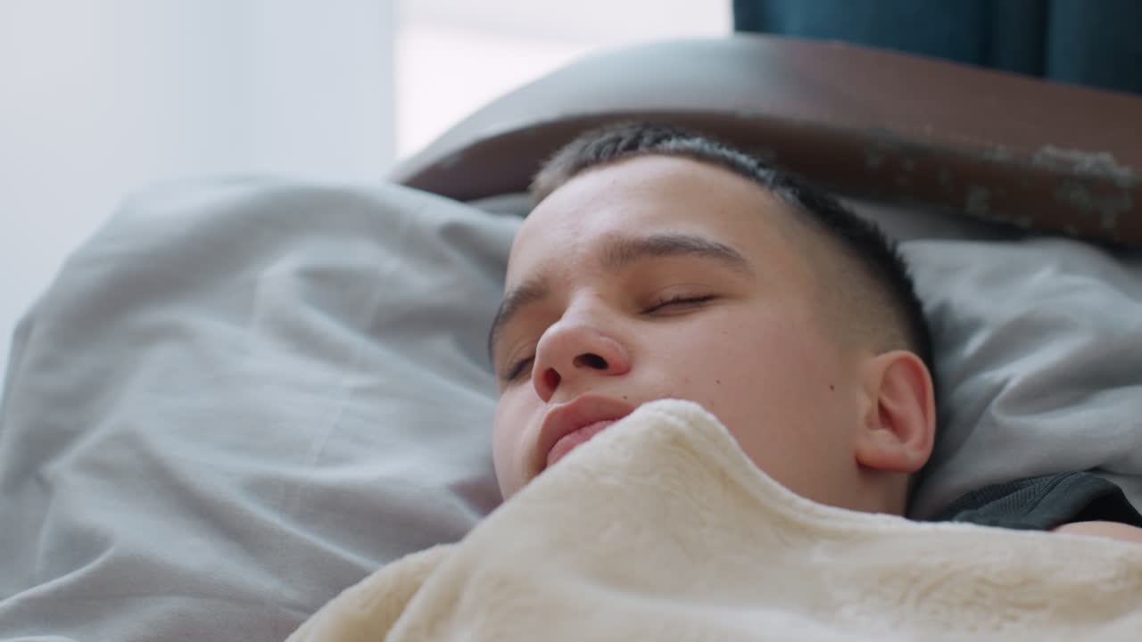 Close up of child yawning and stretching under beige blanket while opening eyes with warm smile in cozy bedroom filled with natural daylight showing relaxed morning wakeup