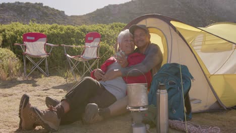 Happy-senior-biracial-couple-sitting-at-tent-in-mountains-and-embracing-on-sunny-day,-in-slow-motion