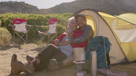 happy senior biracial couple sitting at tent in mountains and embracing on sunny day, in slow motion