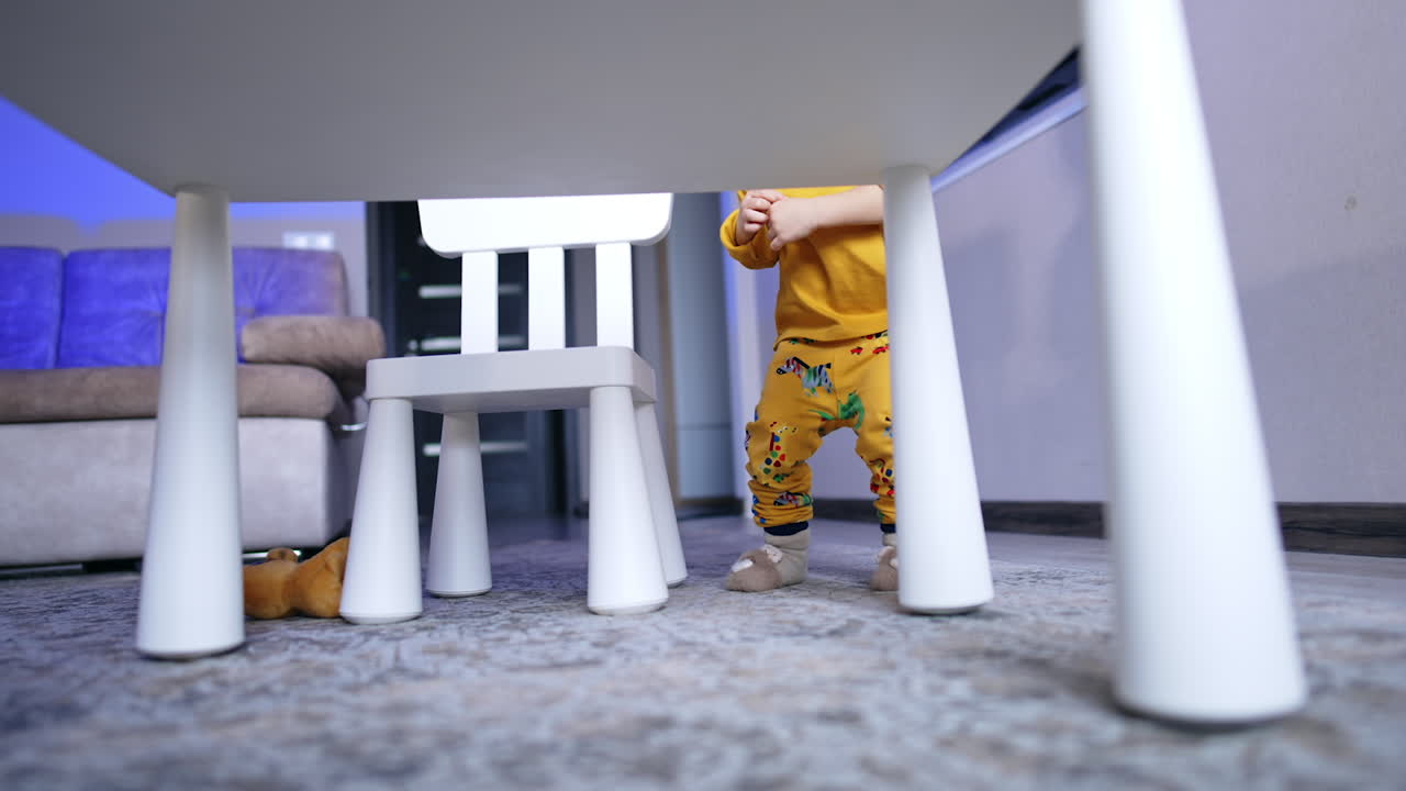 Lovely Caucasian toddler sitting squatted at the desk. Kid in yellow clothes stands up and runs up to camera.