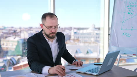 portrait of a serious office worker indoors. businessman in glasses sitting at desk and working on new project in office near the window.