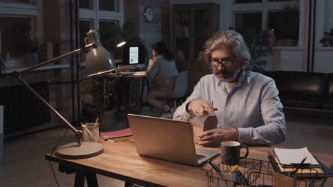 man working late on laptop and eating takeout noodles