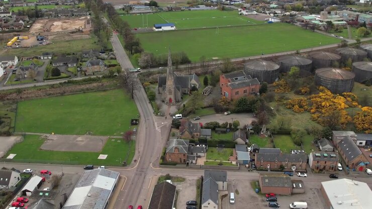 Veduta aerea del centro di Invergordon con edifici storici, una chiesa e serbatoi di stoccaggio industriali circondati da una vegetazione rigogliosa. Cattura elementi urbani e industriali