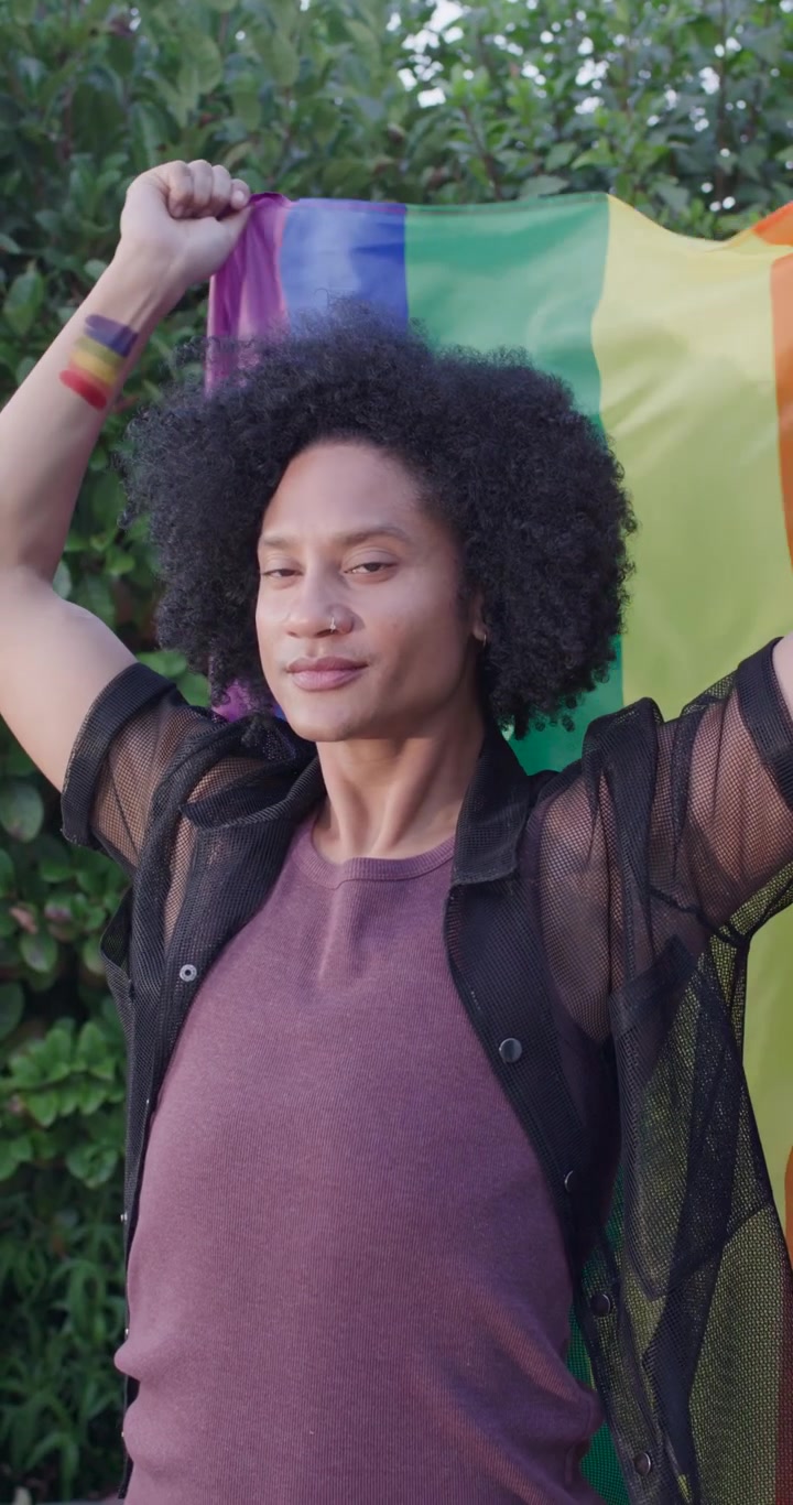 Vídeo vertical: Homem afro-americano levantando a bandeira do arco-íris, ajustando-a e mostrando orgulho ao ar livre
