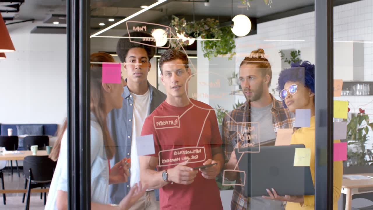 felices colegas creativos diversos haciendo lluvia de ideas con su portátil tomando notas en la pared de vidrio, cámara lenta