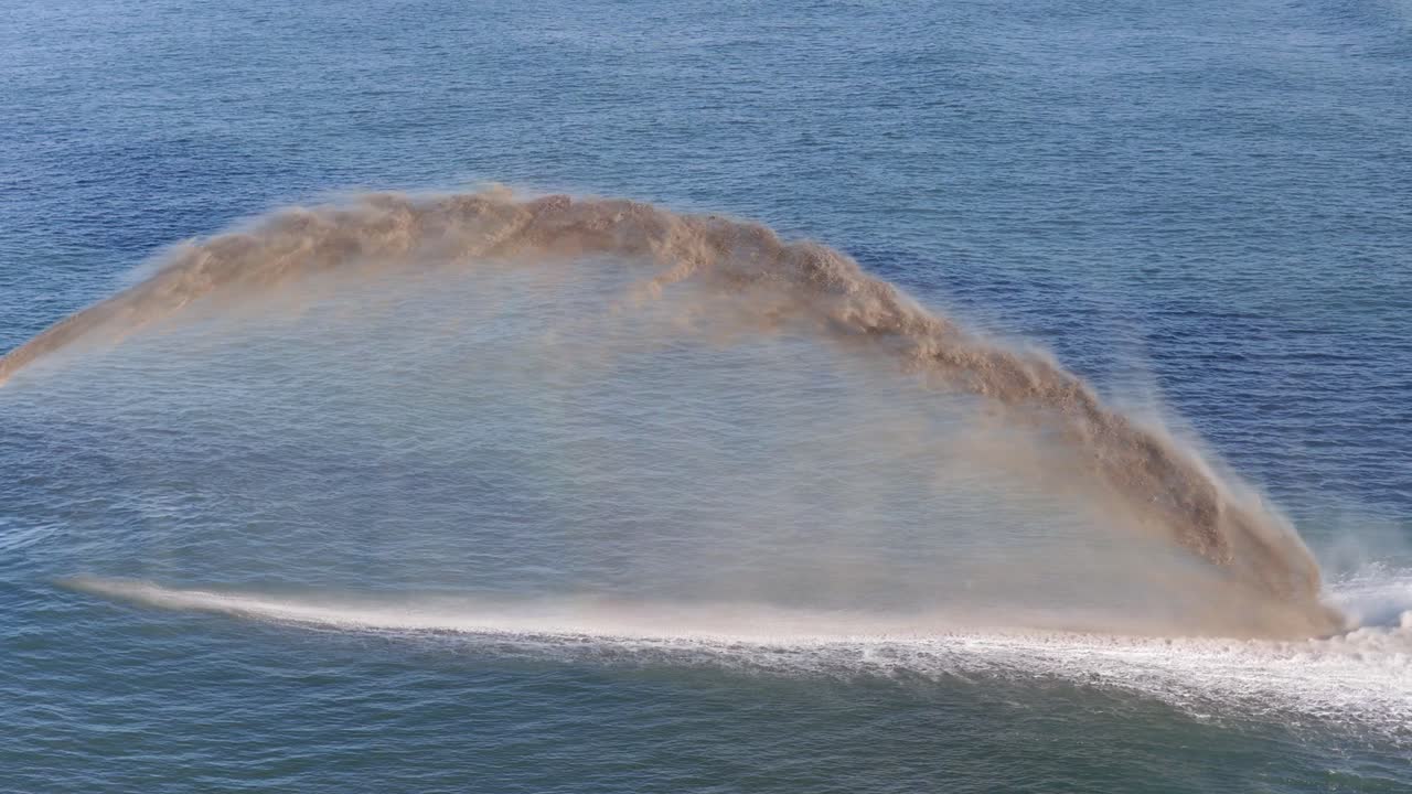Aerial view of a dredger spraying sand into the ocean, creating dynamic patterns against the blue water
