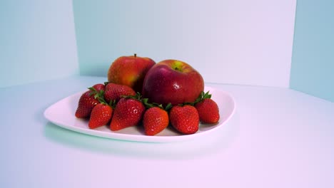 fresh big red tasty ripe strawberries and red apples covered by water droplets rotates slowly on a white plate on light blue background, healthy food concept, medium shot, camera rotate right