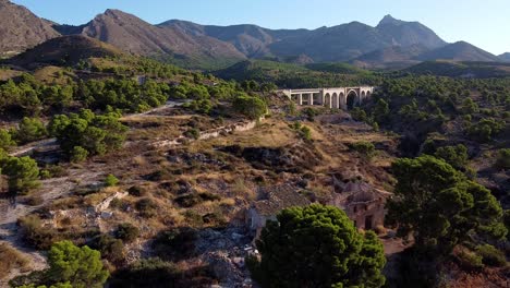 Flying-over-the-ruins-of-an-old-stone-house,-with-an-arched-bridge-in-the-background