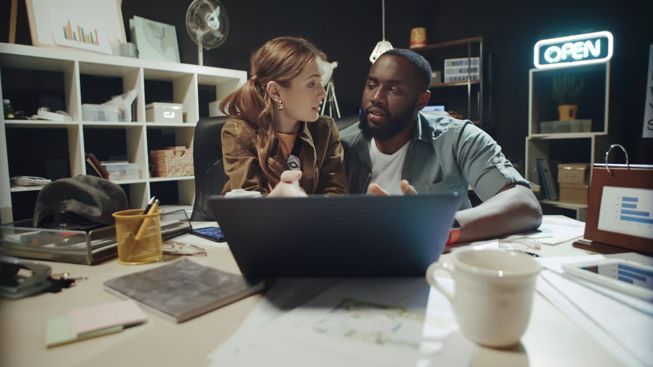 Friendly girl talking to afro guy about data on laptop screen in workplace.